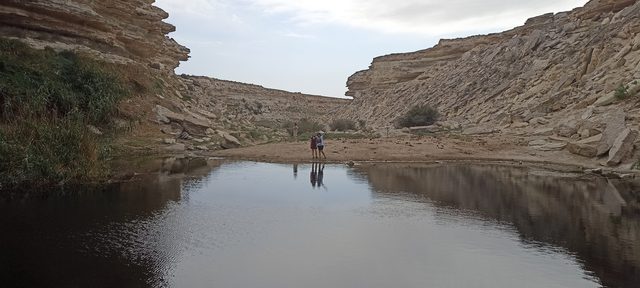 Двое гостей у кромки озера Саура — отражение фигур в спокойной воде между слоистыми скалами.
