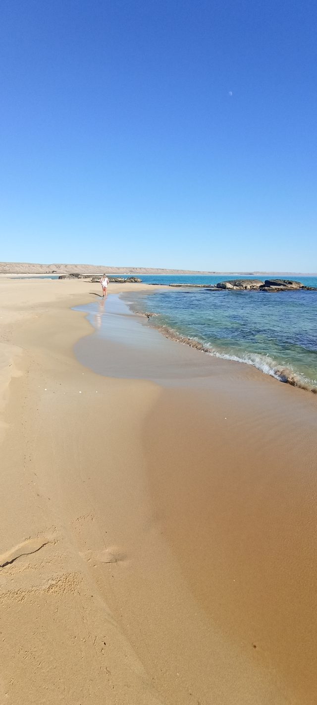 Песчаный пляж у Саура: человек идёт по кромке прибоя, вода прозрачная до самого дна.