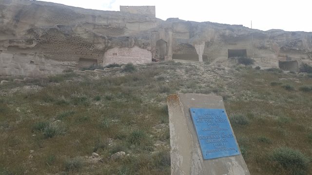Общий вид Шакпак-Ата со склона: мечеть врезана в изъеденную ветром скалу, внизу памятная табличка.