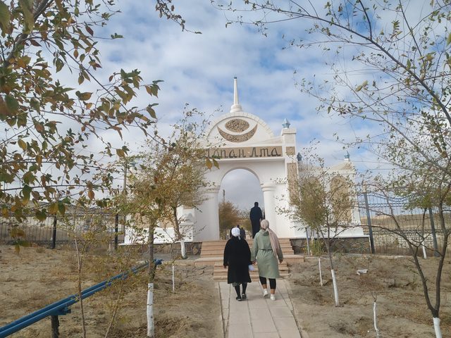 Белые ворота комплекса Шопан-Ата: паломники идут по дорожке к арке между деревьями.
